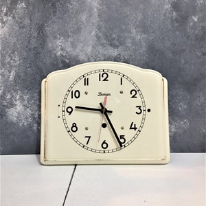 Può includere: Un orologio da parete vintage bianco con lancette nere e numeri romani neri. Il quadrante dell'orologio ha un bordo nero e la parola "Hemingway" stampata in rosa.