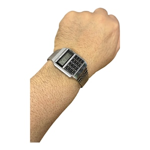 May include: Silver calculator watch with a black keypad and digital display. The watch has a mesh band and is worn on a wrist. The display shows the time. The watch is a vintage style.