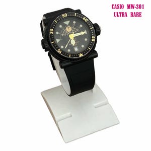 May include: Black Casio MW-301 Ultra Rare watch with a black band and a black face with gold accents. The watch is displayed on a white stand. The watch face has a date display and a compass design.