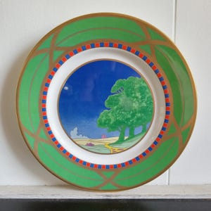 May include: A green and gold decorative plate with a blue and green landscape scene in the center. The scene features two trees and a winding path.