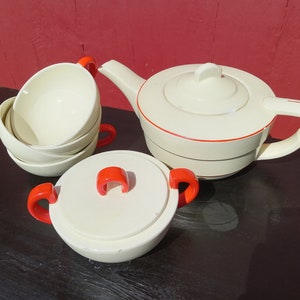 May include: A vintage tea set with a cream-colored teapot, sugar bowl, and three cups. The set has orange accents on the handles and rims.