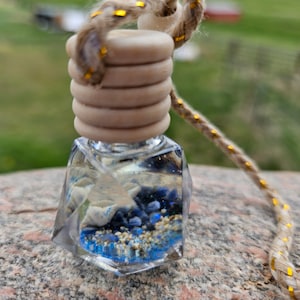Pode incluir: Uma garrafa de vidro transparente com uma rolha de madeira e um cordão dourado. A garrafa está cheia de glitter azul e branco e areia.