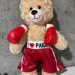 May include: A beige teddy bear wearing red boxing gloves and red boxing shorts with white trim. The shorts have a white heart with the text "I love Paige" on them.