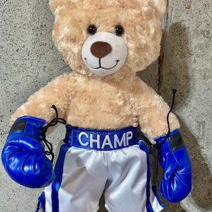 Pode incluir: Um urso de pelúcia com luvas de boxe e calções. O urso é castanho claro com um rosto branco e um nariz castanho. Os calções são brancos com detalhes azuis, e a palavra "CHAMP" está impressa na cintura. As luvas são azuis.