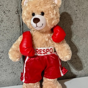 Può includere: Un orsacchiotto di peluche beige con guantoni da boxe rossi e pantaloncini da boxe rossi con una cintura bianca "RESPO".