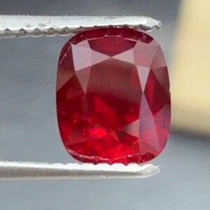 May include: A vibrant, cushion-cut ruby gemstone held by tweezers. The ruby displays a deep red hue with facets that catch the light. The gemstone is a focal point, showcasing its clarity and color. The tweezers are silver-colored.
