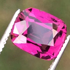 May include: A vibrant, cushion-cut magenta gemstone held by silver-toned metal tweezers. The stone features a faceted surface that reflects light, showcasing deep pink and purple hues throughout its transparent, rectangular-shaped crystalline structure.
