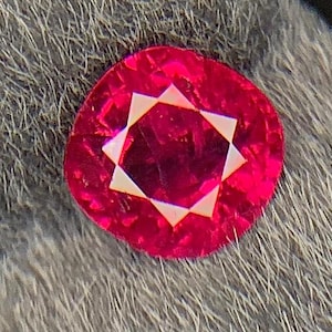 Può includere: Una gemma vibrante, taglio a cuscino, di colore rosso intenso. La gemma presenta un motivo a stella all'interno delle sue sfaccettature, su uno sfondo grigio strutturato. La forma della pietra è un quadrato arrotondato.