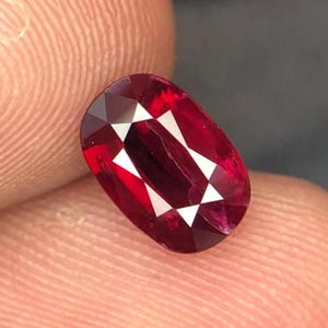 May include: A faceted, oval-shaped ruby gemstone. The gem is a deep red color with white, triangular facets creating a starburst pattern in the center. The ruby is held between two fingers, with a dark background.