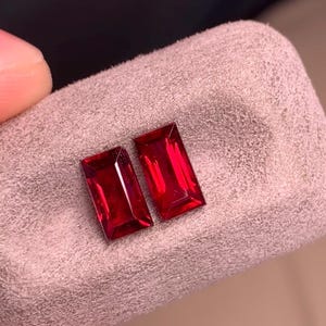 May include: Two rectangular, step-cut, deep red gemstones with a faceted surface. The stones are positioned side-by-side, displaying a vibrant crimson hue and clear internal reflections, showcasing a classic baguette shape with sharp, clean edges.
