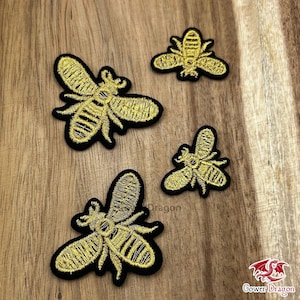 May include: Four embroidered bee patches in different sizes. The bees are gold with black borders, featuring intricate stitching. The patches are displayed on a wooden surface, providing a natural contrast to the vibrant colours of the patches.