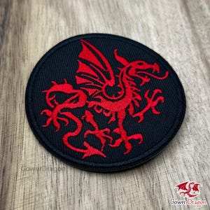 May include: Circular black patch featuring a red dragon emblem. The dragon is intricately embroidered with wings, claws, and a winding body. The patch includes the text "Gower Dragon" at the bottom.