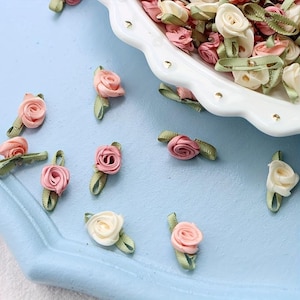 Può includere: Primo piano di rose in miniatura in nastro rosa e crema, con foglie in nastro verde. Alcune rose sono sparse su una superficie azzurra, altre in un piatto bianco con bordo smerlato. Questi delicati abbellimenti floreali sono ideali per lavori creativi.