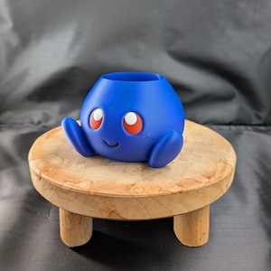 May include: A blue, spherical planter with a smooth finish, featuring two small, rounded side appendages, a wide circular opening at the top, and a face with two large, red-rimmed white eyes and a small, simple, curved black line for a mouth.