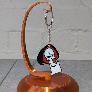 May include: A copper-colored display stand holding a keychain. The keychain features a cartoon grim reaper design with a black cloak, red hood, and white skull. The stand has a circular base and a curved arm.
