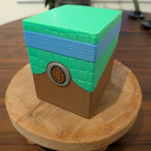 May include: A cubic container featuring a brown base, a green middle section with a grid pattern, and a light blue horizontal band. A circular emblem with a brown hexagonal pattern sits on the front, framed by a grey border. The object has a flat top.