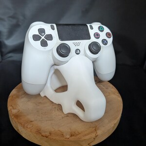 May include: A white wireless video game controller with black analog sticks and a black touchpad, resting on a white, skull-shaped display stand. The controller features standard directional buttons and four colored action symbols.