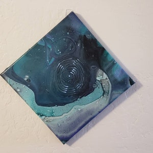May include: Abstract painting on canvas with swirling blue, green, and white paint. The painting has a dark blue background with a white spiral in the center.