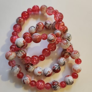 May include: Three beaded bracelets. The bracelets feature a mix of red and white beads. Some beads are solid red, while others are white with red and black abstract designs. The bracelets are elastic and appear to be made of glass or acrylic.