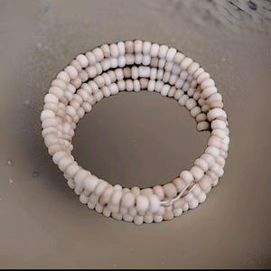 May include: A handmade bracelet composed of small, off-white beads. The beads are strung together in multiple rows, creating a layered effect. The bracelet is secured with a silver-colored clasp. The overall design is simple and elegant.