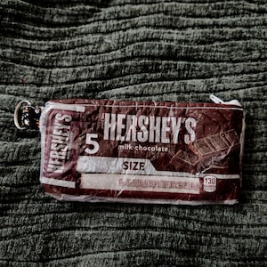 May include: A brown Hershey's milk chocolate-themed zippered pouch. The pouch features the Hershey's logo and the words "5 SIZE" printed on the front. The pouch has a silver-colored key ring attached to the zipper.