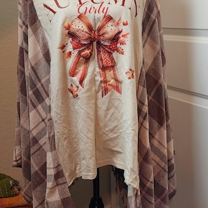 May include: A cream-colored long-sleeve shirt featuring the text AUTUMN Girly and a large red and brown patterned bow graphic surrounded by autumn leaves. The shirt has brown and tan plaid fabric panels on the sides and fringe at the hem.
