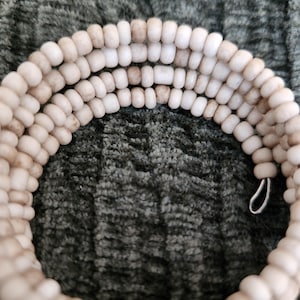 May include: A coiled bracelet made of small, off-white, cylindrical beads. The beads are strung together, forming multiple rows. The bracelet has a silver-colored clasp. The background is a textured, dark green fabric.