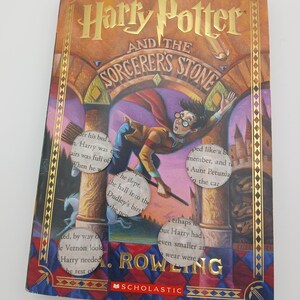 Puede incluir: Un libro de tapa dura titulado "Harry Potter y la piedra filosofal" de J.K. Rowling. La portada dorada presenta el título, una ilustración de Harry Potter en una escoba y un borde decorativo.