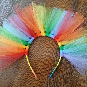 Puede incluir: Una diadema de tul con los colores del arcoíris. La diadema presenta un espectro de colores, incluyendo rojo, naranja, amarillo, verde, azul y morado. El tul está unido a una diadema amarilla.