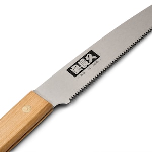 May include: A close-up of a small, folding saw with a wooden handle. The blade is silver and has a serrated edge. The saw is labeled "Silky" in Japanese characters and "Made in Japan".