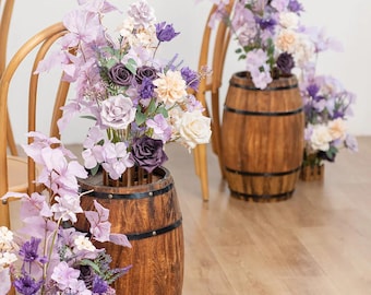 May include: Two wooden barrels with purple and white flowers arranged in them. The barrels are placed in front of wooden chairs.