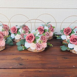 May include: Six centerpieces with a rustic theme. Each centerpiece features a wooden slice base with a floral arrangement of blush pink and white roses, greenery, and small white flowers. A gold metal hoop encircles each arrangement.