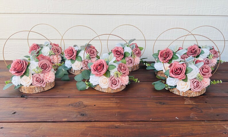 May include: Six centerpieces with a rustic theme. Each centerpiece features a wooden slice base with a floral arrangement of blush pink and white roses, greenery, and small white flowers. A gold metal hoop encircles each arrangement.