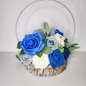May include: A floral arrangement featuring blue, white, and gray roses, accented with greenery. The flowers are arranged on a wooden base and encircled by a silver metal hoop. A decorative piece for weddings or home decor.