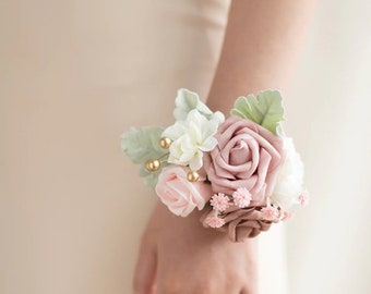 May include: A pink and white flower wrist corsage with green leaves and gold accents.