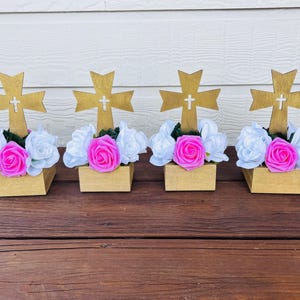May include: Four decorative arrangements featuring gold-colored crosses with white crosses in the center. Each cross is set in a small, square, wooden box filled with white and pink flowers. The arrangement is set on a brown wooden surface.