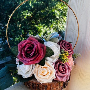 May include: A wooden slice with a gold metal ring, decorated with a bouquet of artificial flowers. The flowers are in shades of burgundy, white, and cream, with green leaves and small yellow berries.