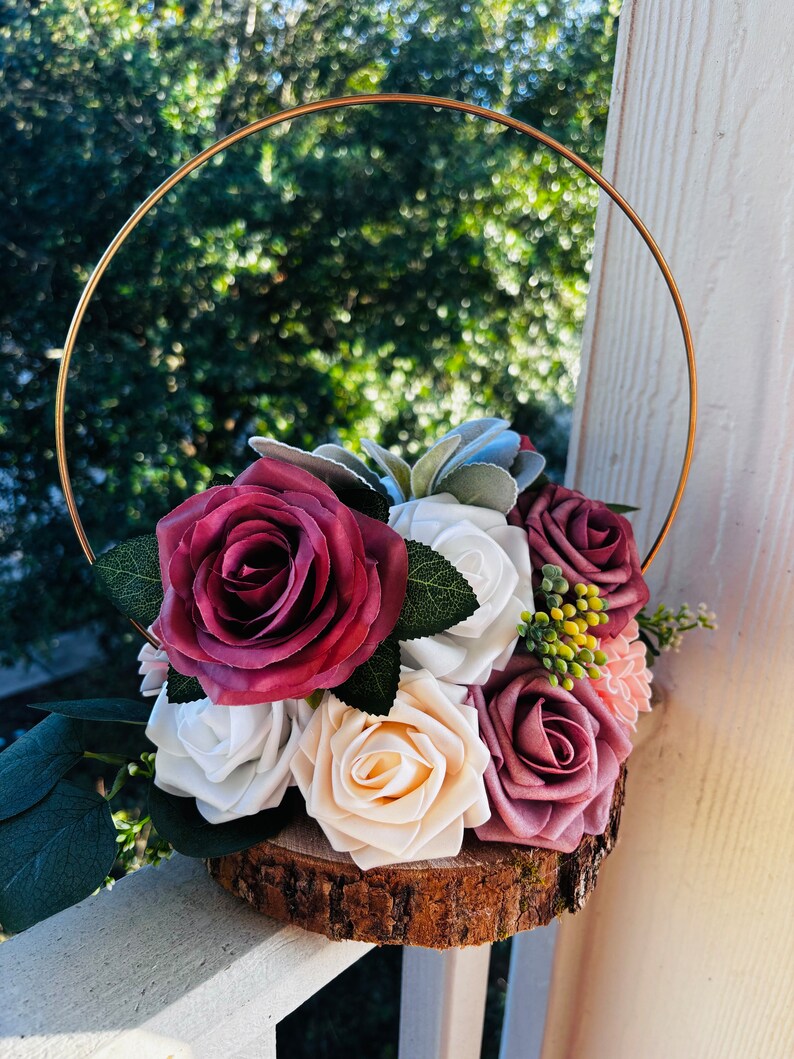 May include: A wooden slice with a gold metal ring, decorated with a bouquet of artificial flowers. The flowers are in shades of burgundy, white, and cream, with green leaves and small yellow berries.