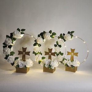 May include: Three white floral wreaths with gold wire hoops and LED lights, each centered around a wooden cross with a white cross design. The wreaths are displayed on wooden bases.