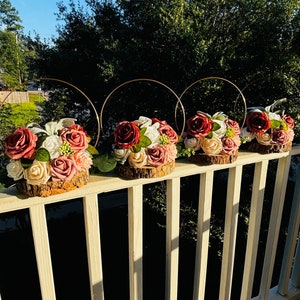 May include: Four floral centerpieces with burgundy and blush roses, greenery, and white flowers arranged on wood slices. Each centerpiece is topped with a gold metal hoop.