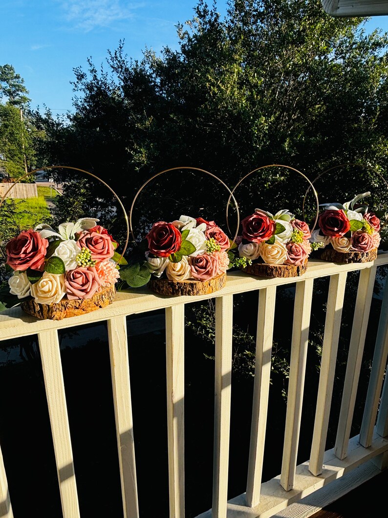 May include: Four floral centerpieces with burgundy and blush roses, greenery, and white flowers arranged on wood slices. Each centerpiece is topped with a gold metal hoop.