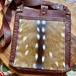 May include: A brown leather crossbody bag with a flap of spotted animal hide. The bag has a long adjustable strap and a stitched border. The bag is made of brown leather and has brass hardware.