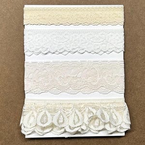May include: A collection of four distinct strips of cream and white lace trim arranged vertically on a white rectangular card. The trims feature various floral and scalloped patterns, ranging from a narrow band to a wider, ruffled edge design.