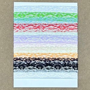 May include: A rectangular white card wrapped with multiple horizontal strips of lace trim in various colors including white, lime green, turquoise, red, lavender, pale yellow, orange, and black, featuring intricate floral patterns throughout.