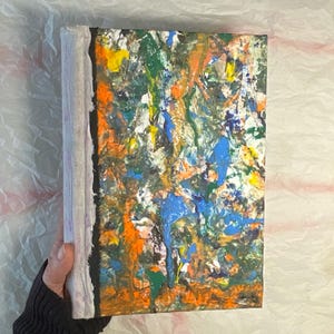 May include: A handmade journal with a colorful abstract painting on the cover. The cover is a mix of blue, green, orange, and white paint splatters. The journal has a white spine and black edges.