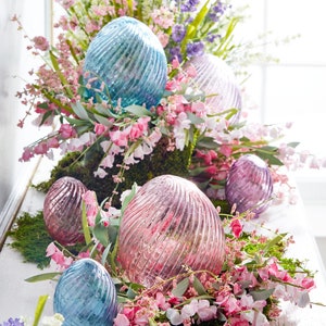 May include: A collection of iridescent glass Easter eggs in shades of blue and pink, nestled in a bed of moss and pink and white flowers.