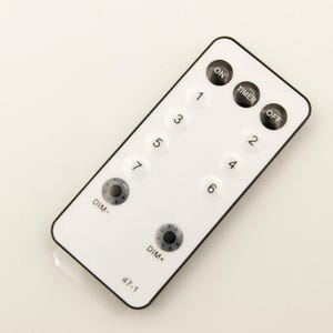 Op de afbeelding: Witte afstandsbediening met zwarte knoppen met de labels "ON", "TIMER", "OFF", "DIM-", "DIM+" en de nummers 1 tot en met 7. De afstandsbediening heeft de tekst "47-1" onderaan.
