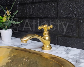 May include: A gold-toned oval sink with a gold-toned faucet with a cross-shaped handle. The sink is on a white countertop with a black brick wall in the background.