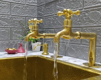 May include: Two gold faucets with handles in the shape of a cross, pouring water into a gold hammered metal sink. The faucets are mounted on a white marble countertop.