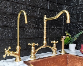 Op de afbeelding: Een gouden keuken spoelbak met een gehamerd koperen afwerking. De spoelbak heeft een enkele kraan met een hoge uitloop en twee kleinere kranen aan beide zijden. De spoelbak is geplaatst tegen een zwarte bakstenen muur.